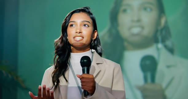A woman in a cream blazer holding a microphone and speaking on stage, with her face shown larger on a screen behind her.