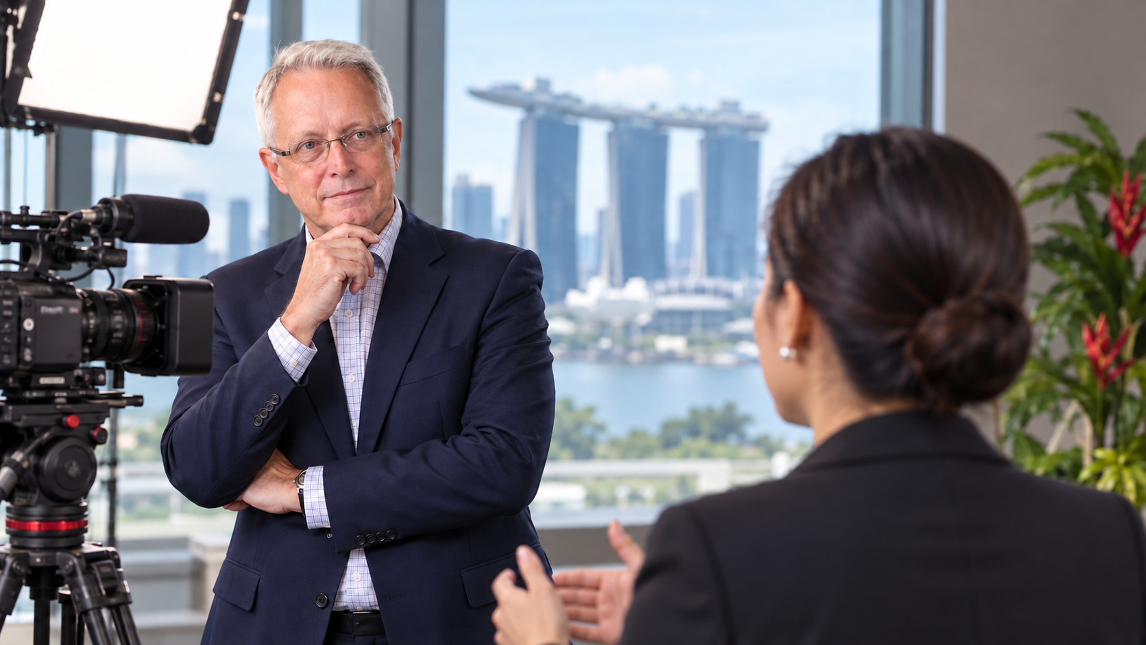 A man in a suit with his hand to his chin, being interviewed by a woman with a camera and studio light beside them, with a city skyline behind.